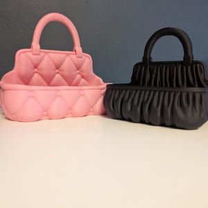 Op de afbeelding: Twee decoratieve handtasvormige containers. E&eacute;n is roze met een gewatteerd ontwerp en de andere is zwart met een geplooid stoffen uiterlijk. Beide hebben een gebogen handvat aan de bovenkant en een rechthoekige basis.