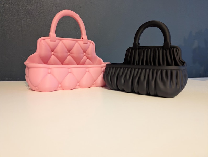 Op de afbeelding: Twee decoratieve handtasvormige containers. E&eacute;n is roze met een gewatteerd ontwerp en de andere is zwart met een geplooid stoffen uiterlijk. Beide hebben een gebogen handvat aan de bovenkant en een rechthoekige basis.