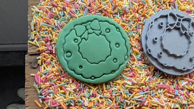 Peut inclure: Un biscuit vert avec un motif de couronne et un emporte-pi&egrave;ce gris avec le m&ecirc;me motif de couronne, le tout sur un lit de vermicelles color&eacute;s. La couronne a un n&oelig;ud en haut et de petits cercles autour.