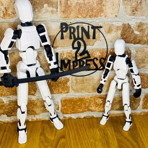 May include: Two white articulated action figures with black joints and accents, one holding a black sword. The figures stand in front of a brick wall with a black sign that reads "PRINT 2 IMPRESS".