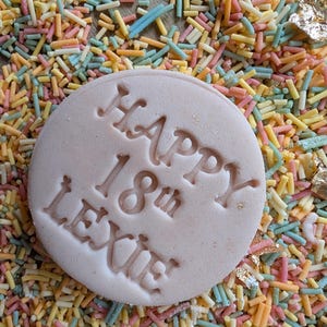 Puede incluir: Una galleta redonda de color rosa claro con las palabras "HAPPY 18th LEXIE" grabadas en la superficie. La galleta est&aacute; rodeada de coloridas virutas en tonos amarillos, rosas, azules y naranjas. Tambi&eacute;n se ven detalles de pan de oro.