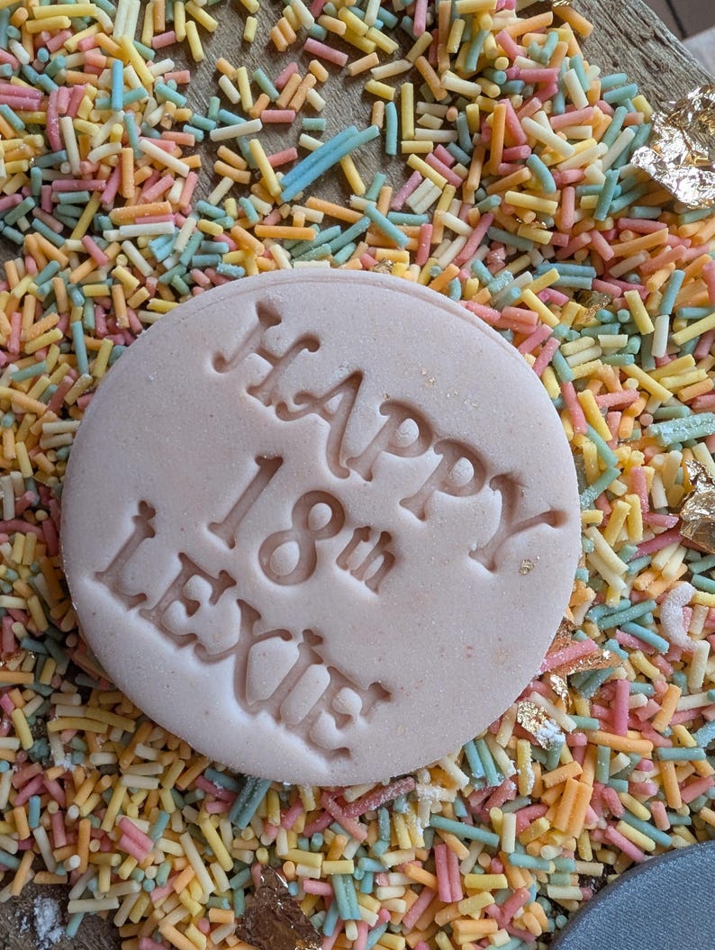 Puede incluir: Una galleta redonda de color rosa claro con las palabras "HAPPY 18th LEXIE" grabadas en la superficie. La galleta est&aacute; rodeada de coloridas virutas en tonos amarillos, rosas, azules y naranjas. Tambi&eacute;n se ven detalles de pan de oro.