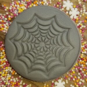 May include: A gray, round, silicone cookie cutter with a raised design of a spider web.