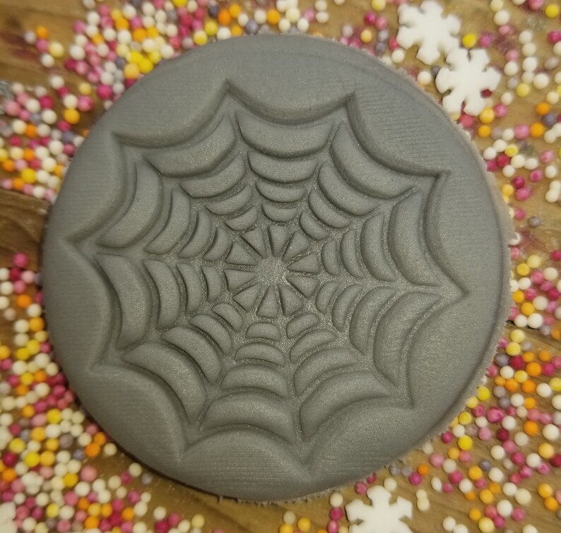 May include: A gray, round, silicone cookie cutter with a raised design of a spider web.