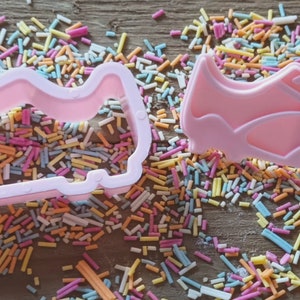 Może przedstawiać: Dwa r&oacute;żowe plastikowe wykrawacze w kształcie but&oacute;w piłkarskich, jeden jako prosty kontur, a drugi bardziej szczeg&oacute;łowy. Otoczone są kolorową posypką na drewnianej powierzchni. Idealne do pieczenia.