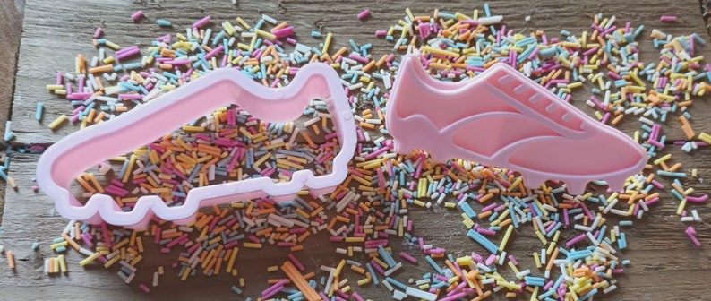 Może przedstawiać: Dwa r&oacute;żowe plastikowe wykrawacze w kształcie but&oacute;w piłkarskich, jeden jako prosty kontur, a drugi bardziej szczeg&oacute;łowy. Otoczone są kolorową posypką na drewnianej powierzchni. Idealne do pieczenia.