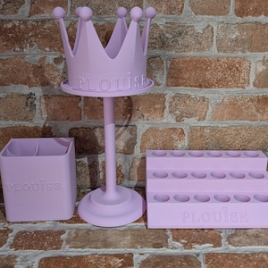 May include: A set of three light purple organizers. The set includes a crown-shaped stand, a two-compartment square container, and a tiered tray with multiple circular openings. The word "PLouise" is printed on each item.