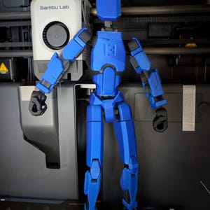 Pu&ograve; includere: Una figura d'azione blu articolata con giunti neri &egrave; in piedi davanti a una stampante 3D. La figura ha un design a blocchi con cerniere visibili e il numero "13" sul petto. La stampante 3D ha il logo "Bambu Lab".
