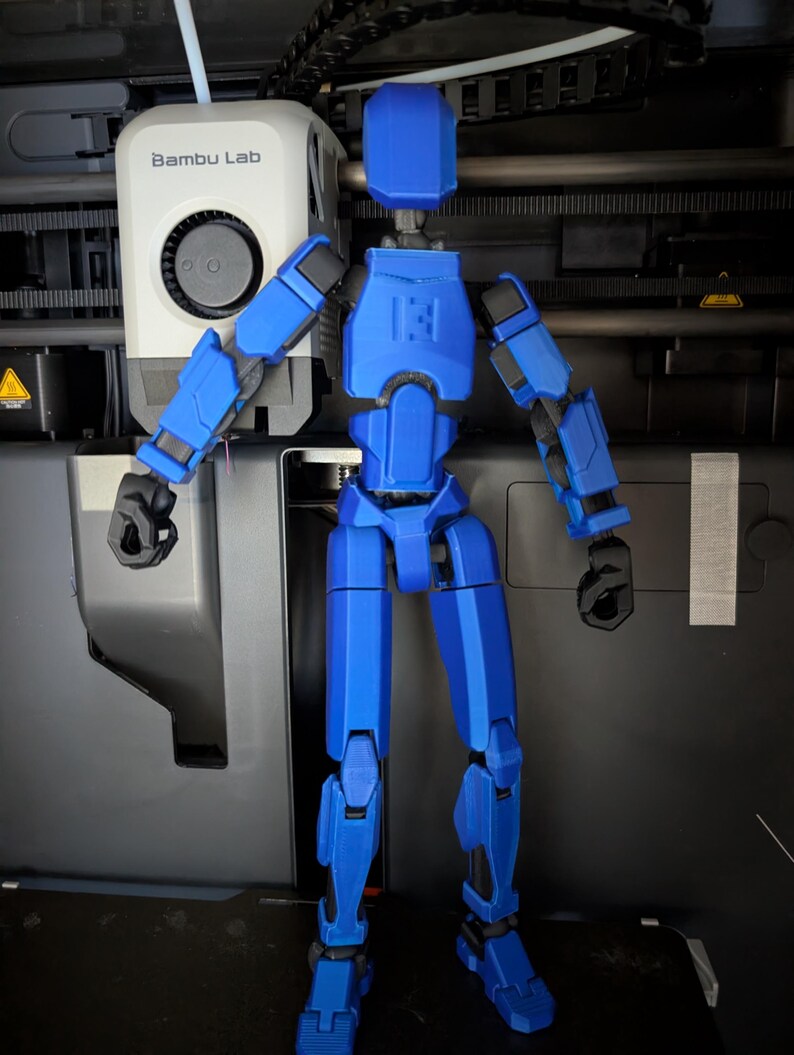 Pu&ograve; includere: Una figura d'azione blu articolata con giunti neri &egrave; in piedi davanti a una stampante 3D. La figura ha un design a blocchi con cerniere visibili e il numero "13" sul petto. La stampante 3D ha il logo "Bambu Lab".