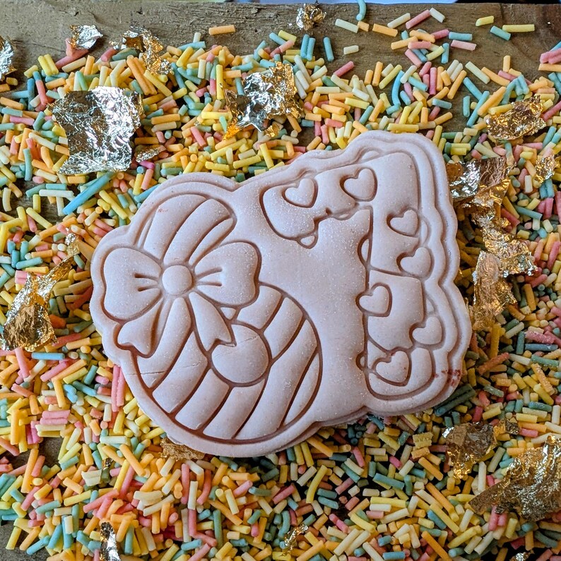 May include: A light pink cookie cutter in the shape of a decorated Easter egg with a bow and hearts. The cookie cutter is surrounded by colorful sprinkles and gold leaf flakes.