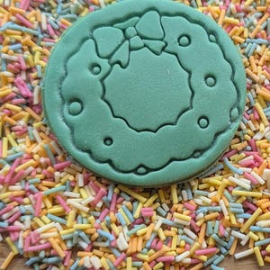 Peut inclure: Un emporte-pi&egrave;ce vert, de forme circulaire, repr&eacute;sentant une couronne de No&euml;l avec un n&oelig;ud, est pos&eacute; sur un lit de vermicelles color&eacute;s. Les vermicelles sont un m&eacute;lange de couleurs pastel, dont le rose, le jaune, le bleu et l'orange.