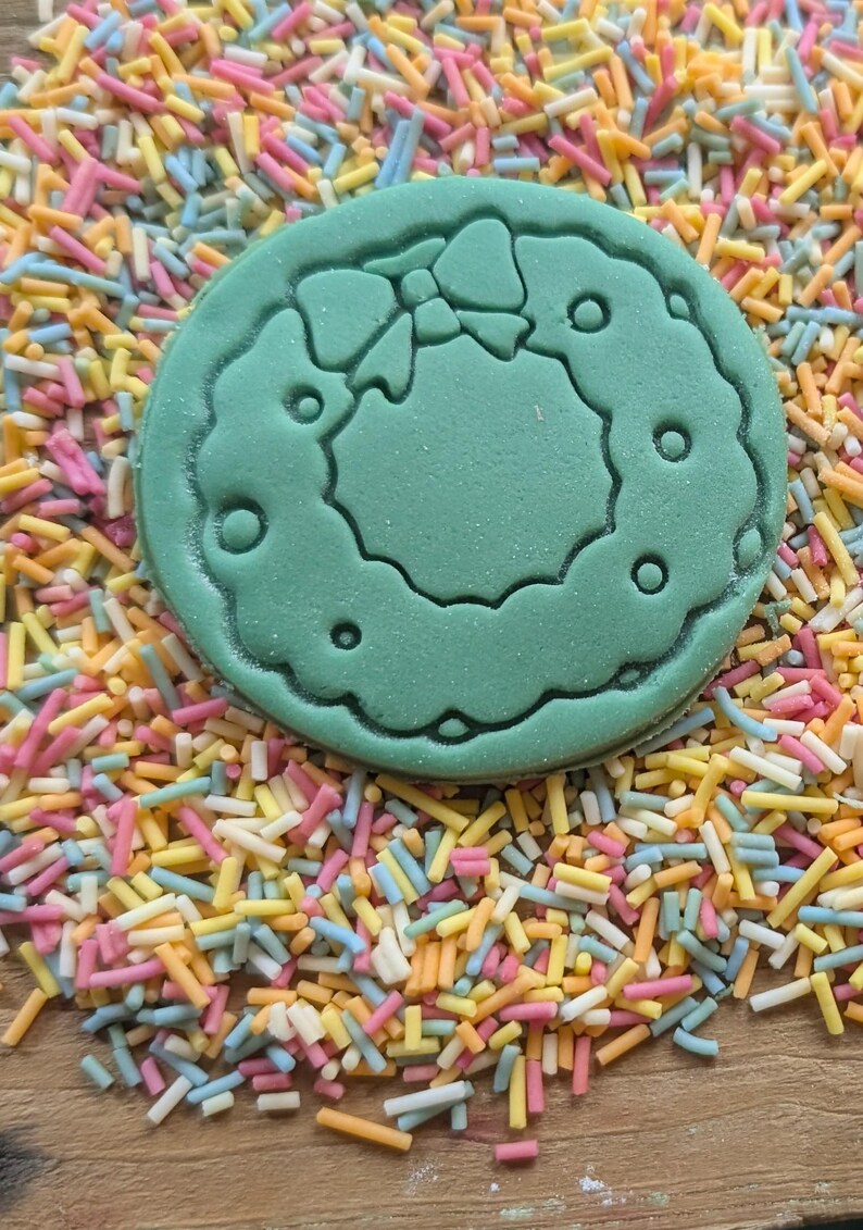 Peut inclure: Un emporte-pi&egrave;ce vert, de forme circulaire, repr&eacute;sentant une couronne de No&euml;l avec un n&oelig;ud, est pos&eacute; sur un lit de vermicelles color&eacute;s. Les vermicelles sont un m&eacute;lange de couleurs pastel, dont le rose, le jaune, le bleu et l'orange.