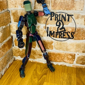 May include: A multi-colored articulated figure with a green head and torso, and limbs in shades of blue, red, and brown. The figure is posed in a dynamic stance, with one arm raised. A black metal sign with the text "PRINT 2 IMPRESS" is in the background.