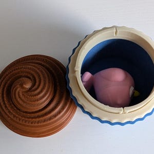 May include: A 3D-printed cupcake-shaped container with a brown swirl lid and a blue interior. The container holds a pink bird-like figure with a yellow beak. The container is open, revealing the figure inside.