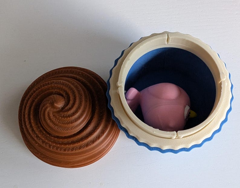 May include: A 3D-printed cupcake-shaped container with a brown swirl lid and a blue interior. The container holds a pink bird-like figure with a yellow beak. The container is open, revealing the figure inside.