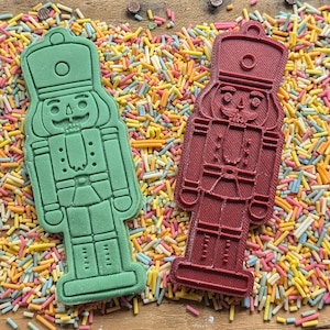 May include: Two nutcracker-shaped cookie cutters, one green and one burgundy, rest on a bed of colorful sprinkles. The cutters have detailed features, including hats, uniforms, and facial expressions. The background is a wooden surface.