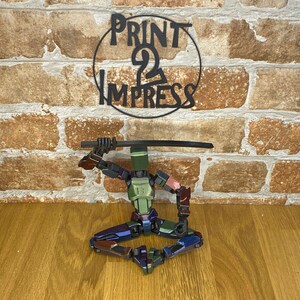 May include: A multi-coloured, articulated robot figure in a seated pose, holding a black sword. The robot has green, blue, and red accents. Above the robot is a black metal sign that reads "PRINT 2 IMPRESS" in a circular design.