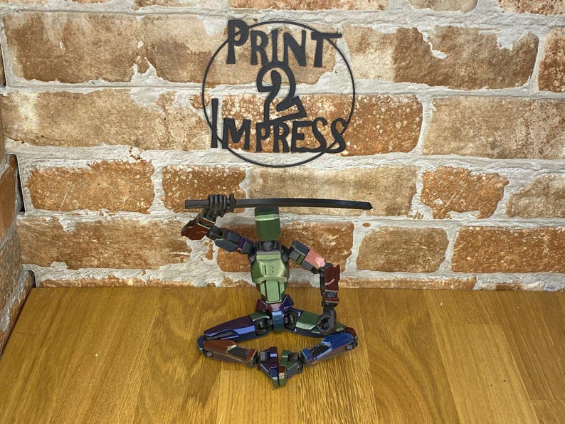 May include: A multi-coloured, articulated robot figure in a seated pose, holding a black sword. The robot has green, blue, and red accents. Above the robot is a black metal sign that reads "PRINT 2 IMPRESS" in a circular design.