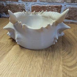 May include: Off-white, crab-shaped decorative bowl with a textured surface and pointed edges. The bowl has a central depression and is placed on a wooden surface. The background features a brick wall.