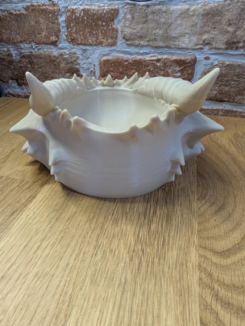 May include: Off-white, crab-shaped decorative bowl with a textured surface and pointed edges. The bowl has a central depression and is placed on a wooden surface. The background features a brick wall.