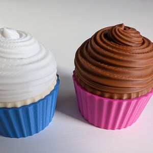 May include: Two decorative cupcake sculptures. One has a white swirl frosting and a blue cupcake liner. The other has a brown swirl frosting and a pink cupcake liner. Both are on a white surface.