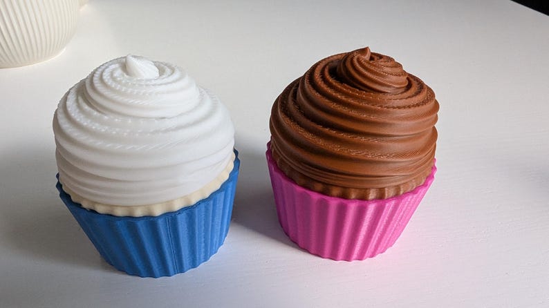 May include: Two decorative cupcake sculptures. One has a white swirl frosting and a blue cupcake liner. The other has a brown swirl frosting and a pink cupcake liner. Both are on a white surface.