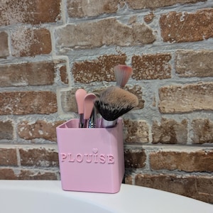 May include: A pink rectangular makeup brush holder with the word "PLouise" embossed on the front, holding several makeup brushes. The brushes have pink, silver, and black handles and various brush heads. The holder sits on a white surface against a brick wall.
