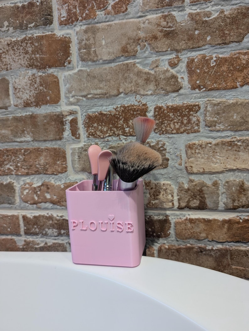 May include: A pink rectangular makeup brush holder with the word "PLouise" embossed on the front, holding several makeup brushes. The brushes have pink, silver, and black handles and various brush heads. The holder sits on a white surface against a brick wall.