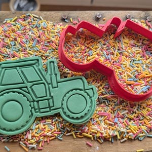 May include: A green tractor-shaped cookie cutter and a red heart-shaped cutter on a wooden surface covered in colourful sprinkles. The tractor cutter is filled with green dough, and the heart cutter is empty.