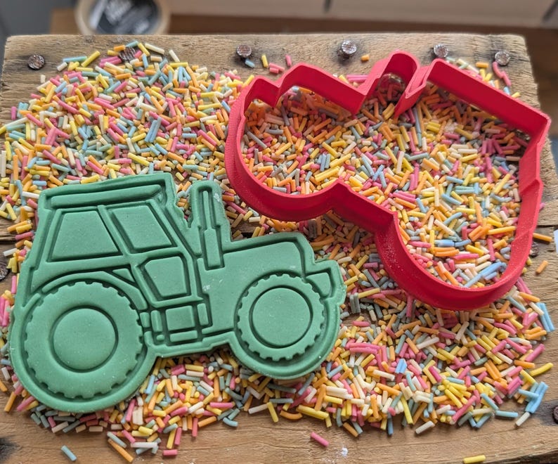 May include: A green tractor-shaped cookie cutter and a red heart-shaped cutter on a wooden surface covered in colourful sprinkles. The tractor cutter is filled with green dough, and the heart cutter is empty.