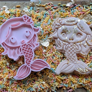 May include: Two pink mermaid cookie cutters with detailed features, including flowing hair and a fishtail. The cutters are surrounded by colorful sprinkles and gold leaf flakes, creating a whimsical scene.