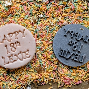 Puede incluir: Dos objetos redondos, uno rosa claro y otro gris, con las palabras "HAPPY 18th LEXIE" en relieve. Est&aacute;n rodeados de fideos de colores y hojuelas de oro.