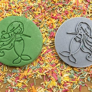 Puede incluir: Dos cortadores de galletas redondos, uno verde y otro gris, con un dise&ntilde;o de sirena. La sirena tiene el pelo largo y fluido y una cola.