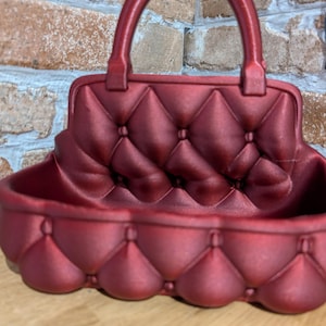May include: A burgundy-colored decorative handbag-shaped planter. The planter features a quilted pattern and a handle. The design resembles a classic handbag, adding a touch of elegance to any space. The planter is made of a solid material.