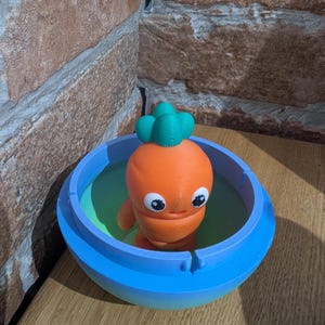 K&ouml;nnte beinhalten: Eine 3D-gedruckte orangefarbene Karottenfigur mit gr&uuml;nem Oberteil in einer blauen Sch&uuml;ssel mit gr&uuml;nem Innenraum. Die Karotte hat gro&szlig;e, runde Augen und kleine Arme. Die Sch&uuml;ssel steht auf einer Holzoberfl&auml;che.