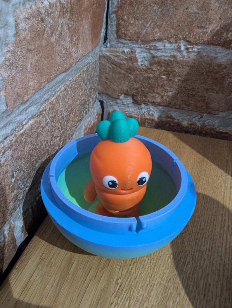 K&ouml;nnte beinhalten: Eine 3D-gedruckte orangefarbene Karottenfigur mit gr&uuml;nem Oberteil in einer blauen Sch&uuml;ssel mit gr&uuml;nem Innenraum. Die Karotte hat gro&szlig;e, runde Augen und kleine Arme. Die Sch&uuml;ssel steht auf einer Holzoberfl&auml;che.
