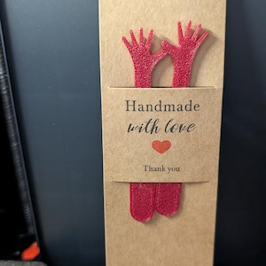Pode incluir: Um marcador de livros feito &agrave; m&atilde;o com fundo castanho. O marcador de livros apresenta duas formas de m&atilde;os vermelhas brilhantes na parte superior e inferior. O centro tem uma etiqueta castanha com as palavras "Handmade with love", um cora&ccedil;&atilde;o vermelho e "Thank you."