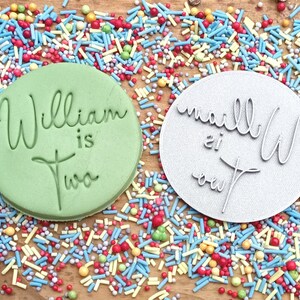 May include: Two round, green and silver cookie cutters with the text "William is Two" and "vanilla is out" on them. The cutters are surrounded by colorful sprinkles.
