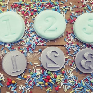 May include: An overhead shot of three light blue circular candies with the numbers 1, 2, and 3 embossed on them. Below are three gray circular objects with the numbers 1, 2, and 3. The candies and objects are surrounded by colorful sprinkles on a wooden surface.