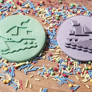 May include: Two round, ship-shaped decorations, one light green and one silver, sit on a wooden surface scattered with colorful sprinkles. The ship design includes sails and waves. A fun, nautical theme.