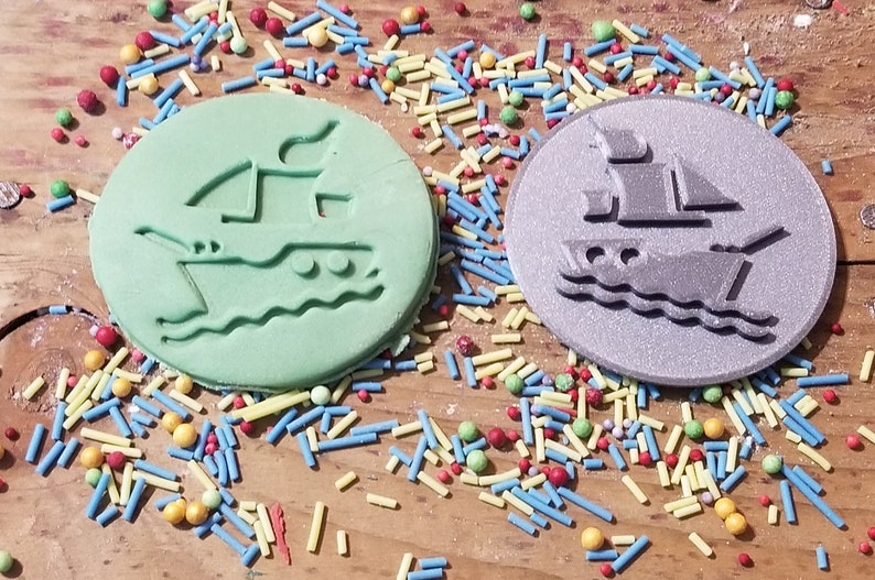 May include: Two round, ship-shaped decorations, one light green and one silver, sit on a wooden surface scattered with colorful sprinkles. The ship design includes sails and waves. A fun, nautical theme.