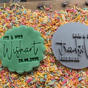 May include: A green, scalloped-edge cookie with the text "Mr. & Mrs. Wishart 28.06.2025" next to a gray circular stamp with the same name and date. The items are surrounded by colorful sprinkles on a wooden surface.