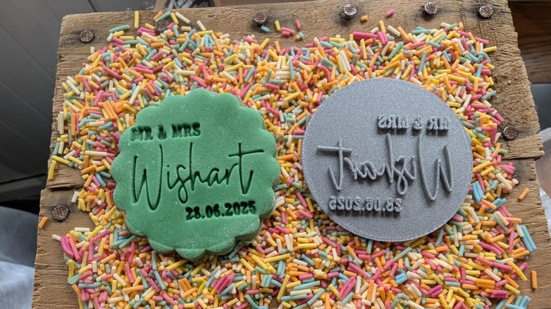 May include: A green, scalloped-edge cookie with the text "Mr. & Mrs. Wishart 28.06.2025" next to a gray circular stamp with the same name and date. The items are surrounded by colorful sprinkles on a wooden surface.