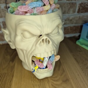 May include: A cream-colored, zombie-head-shaped container filled with colorful, sugared gummy worms. Some gummy worms are also coming out of the mouth. The word "PRINT" is visible in the background.