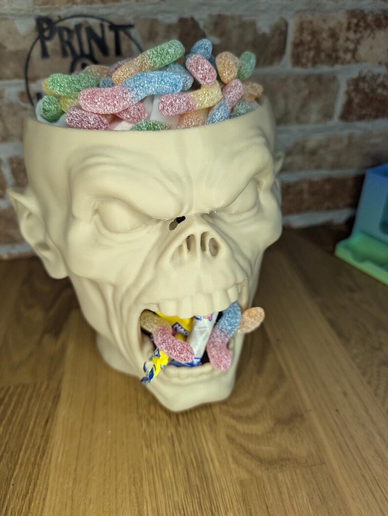 May include: A cream-colored, zombie-head-shaped container filled with colorful, sugared gummy worms. Some gummy worms are also coming out of the mouth. The word "PRINT" is visible in the background.