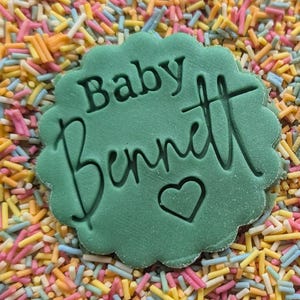 K&ouml;nnte beinhalten: Eine gr&uuml;n gef&auml;rbte Dekoration mit gewelltem Rand, die die Worte "Baby Bennett" und ein Herzsymbol tr&auml;gt. Die Dekoration ist von bunten Streuseln in Rosa, Gelb, Blau und Orange umgeben, was einen festlichen Hintergrund bildet.