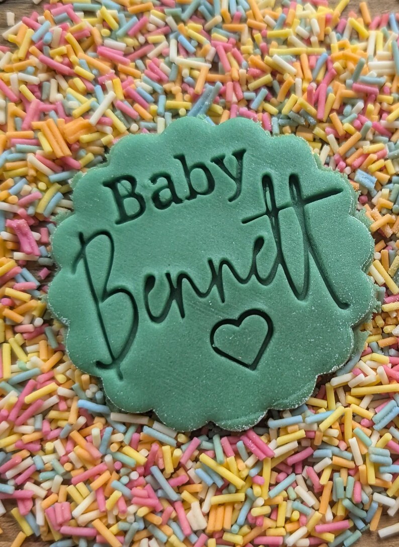 K&ouml;nnte beinhalten: Eine gr&uuml;n gef&auml;rbte Dekoration mit gewelltem Rand, die die Worte "Baby Bennett" und ein Herzsymbol tr&auml;gt. Die Dekoration ist von bunten Streuseln in Rosa, Gelb, Blau und Orange umgeben, was einen festlichen Hintergrund bildet.