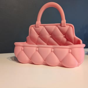 May include: A pink, quilted handbag with a handle. The bag has a rounded base and a rectangular top with a handle. The bag is made of a soft material and has a quilted pattern.