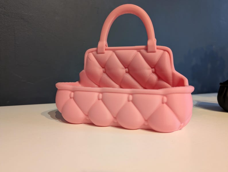 May include: A pink, quilted handbag with a handle. The bag has a rounded base and a rectangular top with a handle. The bag is made of a soft material and has a quilted pattern.