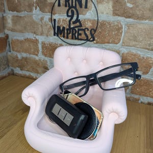 May include: A light pink miniature sofa-shaped organizer holding black-framed eyeglasses, a car key fob, and a small wallet. The organizer is on a wooden surface, with a brick wall in the background. The sign above the sofa reads "PRINT 2 IMPRESS".